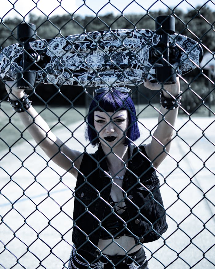 Woman with blue hair holding skateboard behind chain-link fence at skate park.