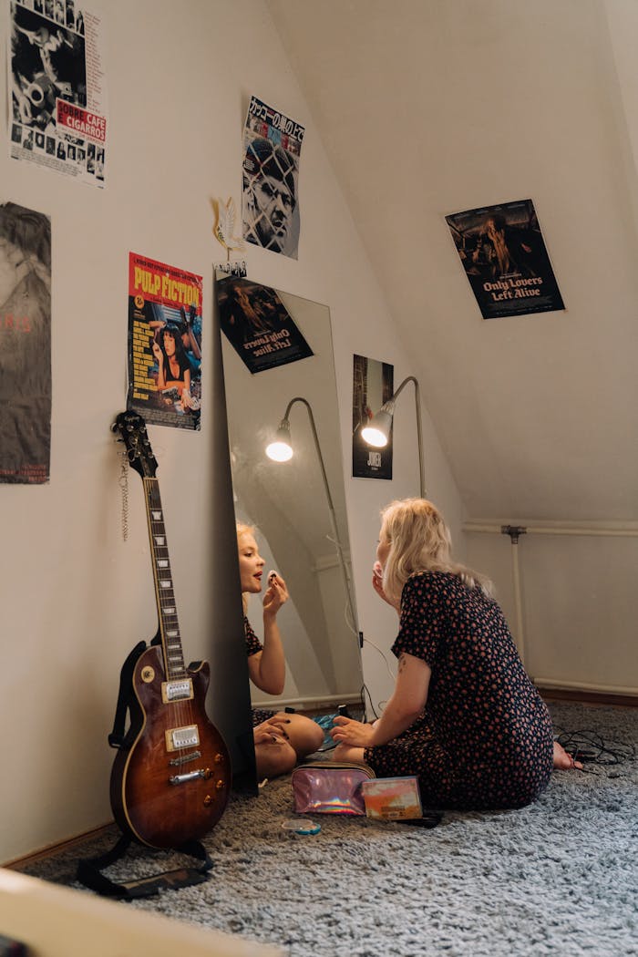 A young Caucasian woman applies makeup in a cozy room with posters and a guitar.